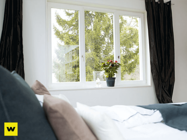 Ein helles Schlafzimmer mit großen Fenstern, Bett und Topfpflanze auf der Fensterbank.